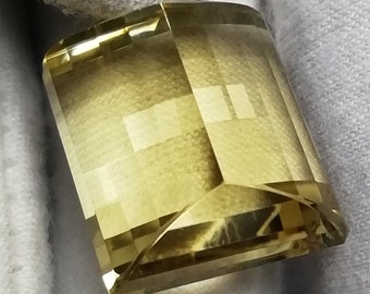 Peut inclure: Une pierre précieuse rectangulaire et facettée, de couleur jaune pâle. La gemme présente de multiples facettes qui réfléchissent la lumière, créant un effet scintillant. La pierre est posée sur un fond blanc.