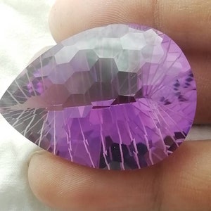 May include: A faceted, teardrop-shaped gemstone with a gradient of purple hues. The gem features intricate cuts that reflect light, creating a sparkling effect. The stone's surface is a deep violet, transitioning to lighter shades.