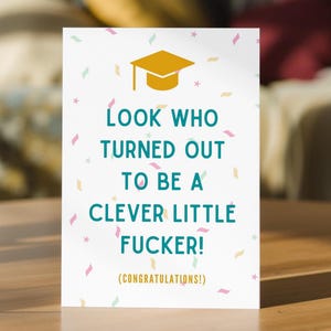 May include: A white greeting card featuring a gold graduation cap icon at the top, teal text that reads "LOOK WHO TURNED OUT TO BE A CLEVER LITTLE FUCKER!" and smaller gold text below stating "(CONGRATULATIONS!)", all surrounded by colourful confetti.
