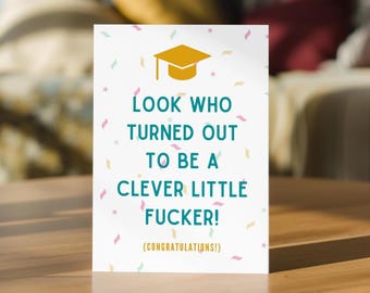 May include: A white greeting card featuring a gold graduation cap icon at the top, teal text that reads "LOOK WHO TURNED OUT TO BE A CLEVER LITTLE FUCKER!" and smaller gold text below stating "(CONGRATULATIONS!)", all surrounded by colorful confetti.