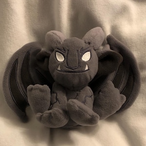 Puede incluir: Un gargoyle de peluche gris con alas, sentado sobre una superficie blanca. El gargoyle tiene una expresión amenazante con ojos blancos y una boca cosida.