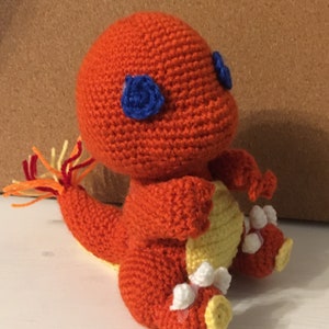 Op de afbeelding: Gehaakt oranje wezen met blauwe ogen, een gele buik en witte en gele accenten op de voeten. De staart heeft oranje, gele en rode garenstrengen. Het handgemaakte pluche speelgoed staat op een wit oppervlak.