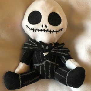 May include: A plush toy of Jack Skellington from the Nightmare Before Christmas. The toy is wearing a black and white striped suit with a black bow tie. The toy has a white face with black eyes, a stitched mouth, and a black nose.