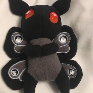 May include: A plush moth toy with a black body, large black wings, and red eyes. The moth has a gray abdomen and wings with a detailed eye design. The toy is made of soft fabric and has a whimsical design.