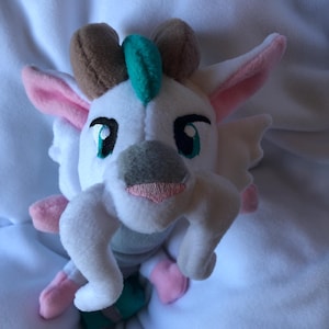 Puede incluir: Un peluche con forma de criatura con detalles en blanco, marrón, rosa y verde. Tiene ojos grandes y expresivos, un hocico gris y una boca rosa. Las orejas están forradas en rosa y tiene brazos y piernas pequeños y curvados. Sobre una superficie blanca.