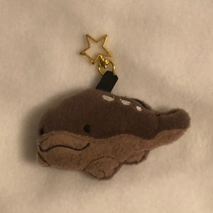 May include: A brown plush toy shaped like a whale with a gold star charm attached. The whale has a small, white, embroidered smile.