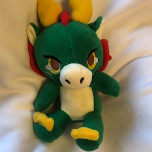 May include: Green plush dragon with yellow horns and red accents. The dragon has a white face with brown eyes and a small, pink nose.