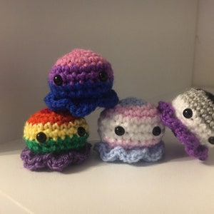 Pode incluir: Quatro medusas de crochê, cada uma com um padrão de cor diferente que representa uma bandeira do orgulho. As medusas são roxas, azuis, brancas e cinzas com detalhes pretos.