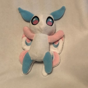 Puede incluir: Un juguete de peluche blanco con alas azules y rosas. El juguete tiene ojos grandes y redondos y una expresión sonriente.