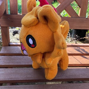 May include: A soft toy creature in shades of orange and red, with large black and white eyes and a pink mouth. The plushie has small ears and is positioned on a wooden bench. The toy is made of a soft material.