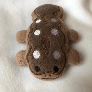 May include: A brown plush toy shaped like a salamander with white and purple embroidered spots. The toy has a stitched-on mouth and eyes.