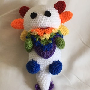 May include: A white crocheted axolotl plush toy with rainbow coloured details. The axolotl has large black eyes and a purple tail.