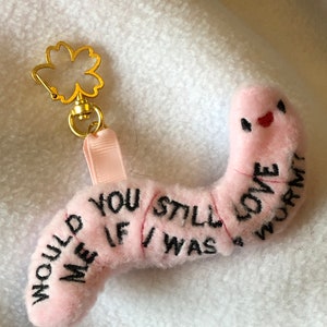 May include: Pink plush worm keyring with the text "Would you still love me if I was a worm?" embroidered on it. The keyring has a gold metal flower charm attached to it.