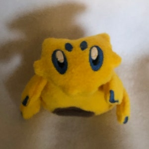 May include: Yellow plush toy with blue eyes and a brown belly. The toy is shaped like a small animal with a round body and a pointed head.