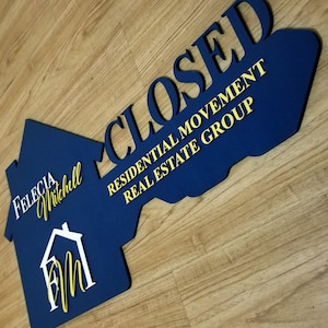 May include: A navy blue key-shaped sign with the word "CLOSED" in bold yellow letters. The sign also includes the text "RESIDENTIAL MOVEMENT REAL ESTATE GROUP" and the name "Felecia Mitchell". A house graphic is also included.
