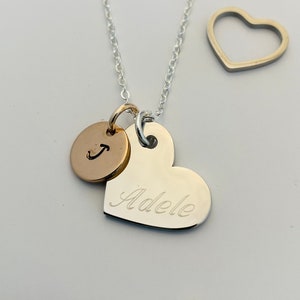 May include: A silver necklace with a silver heart charm engraved with the name "Adele" and a gold circle charm with the letter "J". There is also a separate silver heart charm.