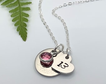May include: A silver necklace with a heart charm engraved with the number "13" and a round charm with a pink gemstone. The necklace is on a white background.