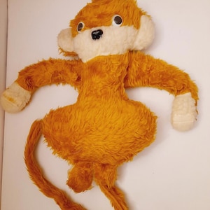 Può includere: Un peluche a forma di scimmia con pelo lungo e arruffato color arancione, viso color crema e mani color crema. Il giocattolo presenta grandi occhi rotondi in plastica bianca e nera, un piccolo naso nero e una lunga coda sottile.