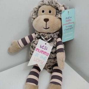 May include: A plush monkey toy with brown and cream striped arms and legs, featuring a fuzzy brown and cream body. A white tag attached to the neck reads "best mummy in the world" in pink and blue text, and a light blue tag reads "Just a little something to say I love you".