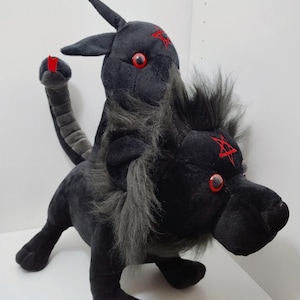 May include: A black plush toy featuring two heads, one resembling a lion with a dark gray mane and the other a horned creature. Both heads have red eyes and a red pentagram embroidered on the forehead. It has a segmented tail with a red tip.