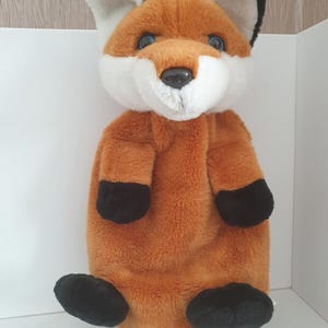 May include: A plush fox toy featuring soft orange fur, a white chest and muzzle, and black accents on the ears and paws. The stuffed animal has large, dark blue eyes and a small black nose, designed with a rounded body and short limbs.