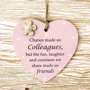 Puede incluir: Placa de madera rosa en forma de coraz&oacute;n con el texto "Chance made us Colleagues, but the fun, laughter and craziness we share made us friends." La placa tiene una peque&ntilde;a flor de madera y lunares blancos. Un lazo de cuerda en la parte superior.