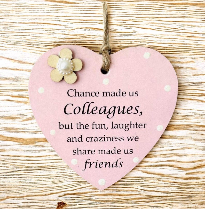 Puede incluir: Placa de madera rosa en forma de coraz&oacute;n con el texto "Chance made us Colleagues, but the fun, laughter and craziness we share made us friends." La placa tiene una peque&ntilde;a flor de madera y lunares blancos. Un lazo de cuerda en la parte superior.