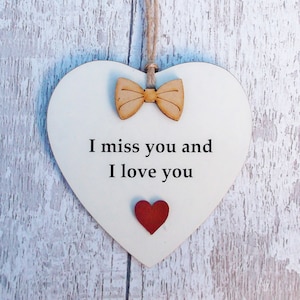Può includere: Un cartello di legno bianco a forma di cuore con il testo "I miss you and I love you" in lettere nere. Un piccolo papillon di legno e un cuore rosso sono attaccati al cartello.