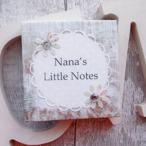 May include: A white wooden box with a lace-trimmed circle and the text "Nana's Little Notes". The circle is decorated with two pink and white flowers with rhinestones in the center.
