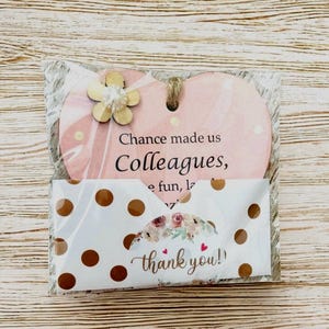 Puede incluir: Un adorno de madera en forma de coraz&oacute;n rosa con el texto "Chance made us Colleagues". Una peque&ntilde;a flor de madera est&aacute; adjunta. Tambi&eacute;n se incluye una tarjeta blanca con lunares dorados y las palabras "thank you!".