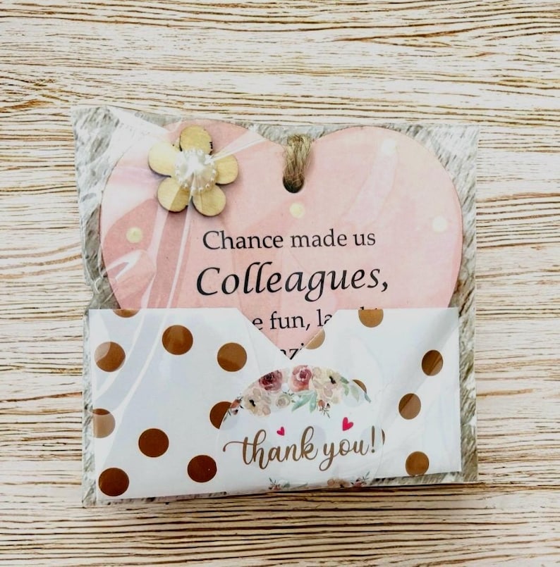 Puede incluir: Un adorno de madera en forma de coraz&oacute;n rosa con el texto "Chance made us Colleagues". Una peque&ntilde;a flor de madera est&aacute; adjunta. Tambi&eacute;n se incluye una tarjeta blanca con lunares dorados y las palabras "thank you!".