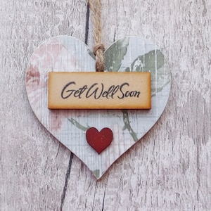 Puede incluir: Un adorno de madera en forma de corazón con un patrón floral y un pequeño corazón rojo. Un letrero de madera con el texto "Get Well Soon" está unido al adorno.