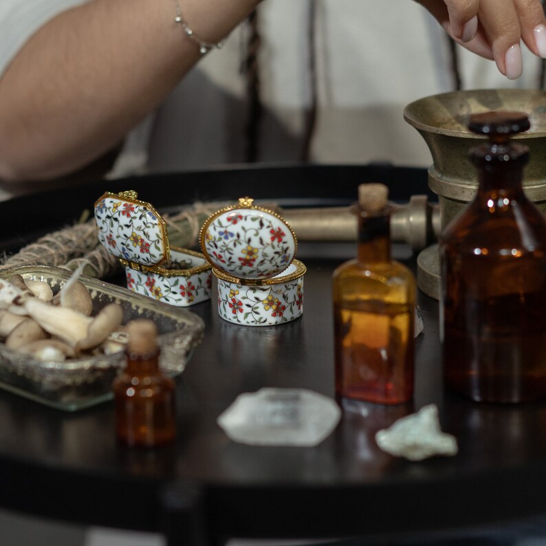 May include: Two small porcelain boxes with floral patterns, a clear glass bottle with a cork stopper, a brown glass bottle, and a small bowl filled with mushrooms on a black tray.
