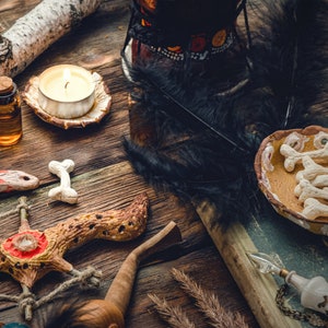 May include: A wooden table with a variety of objects, including a lit candle, a small bottle, a wooden stick, a feather, a small bowl with white ceramic bones, and a wooden sculpture with a red and white design.