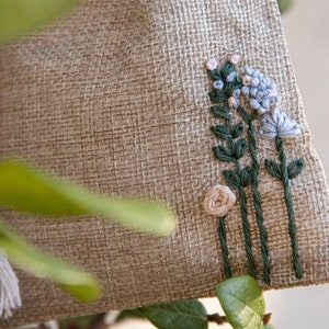 May include: A small, beige burlap bag with a drawstring closure. The bag is decorated with embroidered flowers in shades of pink, blue, and green.