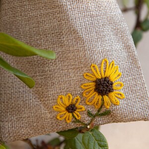 Puede incluir: Una pequeña bolsa de arpillera con dos girasoles bordados. Los girasoles son amarillos con centros marrones y tallos verdes.