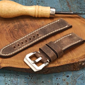 Può includere: Un cinturino in pelle marrone con cuciture bianche e fibbia argentata è appoggiato su una superficie di legno. Sono presenti anche un lesina in legno e un braccialetto di perline. Il cinturino ha un aspetto vintage.