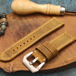 Op de afbeelding: Twee bruinleren horlogebandjes met witte stiksels en een zilveren gesp. Een houten priem en een kralenarmband zijn ook zichtbaar op een houten oppervlak. De bandjes zijn voor een vintage horloge.