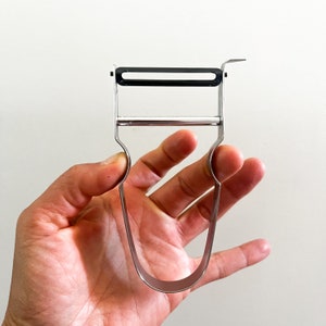 May include: A silver and black peeler with a curved handle. The peeler is designed to peel vegetables and fruits.