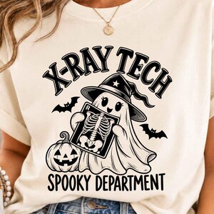 May include: A cream-colored t-shirt featuring a black graphic of a ghost wearing a witch hat, holding an X-ray film of a ribcage. The design includes a jack-o'-lantern, two bats, and the text "X-RAY TECH" and "SPOOKY DEPARTMENT" in black.