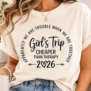 以下が含まれることがあります： クリーム色のTシャツ。黒い文字で「Apparently we are trouble when we are together, Girl's Trip, Cheaper than therapy, 2026」とプリントされており、年号のゼロの部分がハートマークになっていて、両サイドに装飾的な矢印がデザインされています。