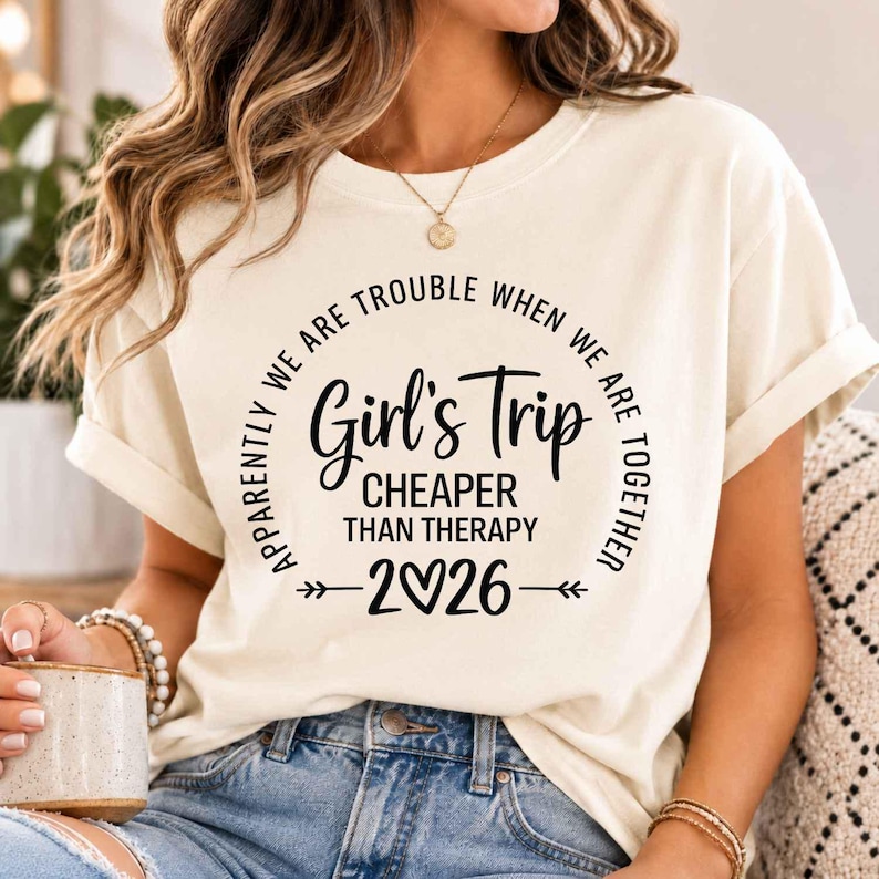 以下が含まれることがあります： クリーム色のTシャツ。黒い文字で「Apparently we are trouble when we are together, Girl's Trip, Cheaper than therapy, 2026」とプリントされており、年号のゼロの部分がハートマークになっていて、両サイドに装飾的な矢印がデザインされています。