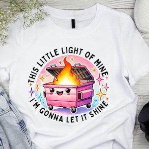 May include: White t-shirt with a pink trash can with a smiling face and flames coming out of it. The text around the trash can says "This little light of mine I'm gonna let it shine."