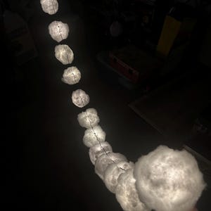 May include: A string of white cloud-shaped lights with a warm glow. The lights are connected by a thin wire and are hanging in a dark room.