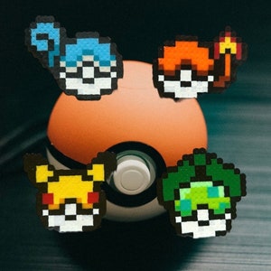 Puede incluir: Primer plano de una Poké Ball con cuatro personajes de Pokémon diferentes a su alrededor. Los personajes están hechos de cuentas Perler de colores e incluyen Bulbasaur, Squirtle, Charmander y Pikachu.