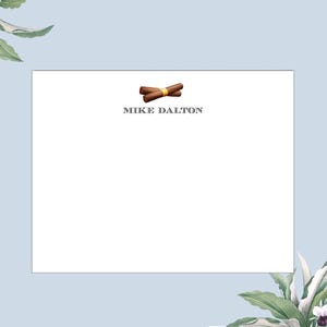 May include: A white card with a brown and yellow illustration of two crossed cigars and the text "MIKE DALTON" printed in black.