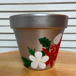 May include: A silver painted terracotta flower pot with a hand-painted design of white and red flowers and green leaves.