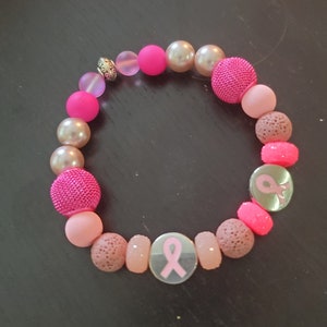 Puede incluir: Pulsera de cuentas rosa y marrón con un dije de cinta rosa. La pulsera está hecha con cuentas de diferentes tamaños y texturas.