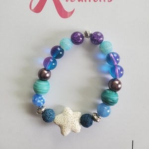 May include: A blue, purple, and green beaded bracelet with a white star charm. The bracelet is made with various types of beads, including lava rock, agate, and glass. The star charm is made of white stone and has a textured surface.