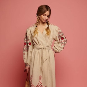 May include: A beige linen dress with red and black embroidered geometric patterns on the sleeves and hem. The dress has a wrap style and a belt.